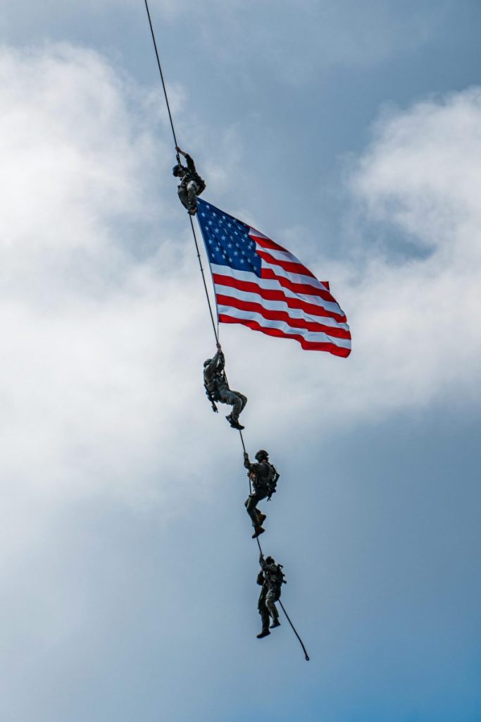 Soldiers conduct a dramatic helicopter rescue holding the American flag against a cloudy sky.